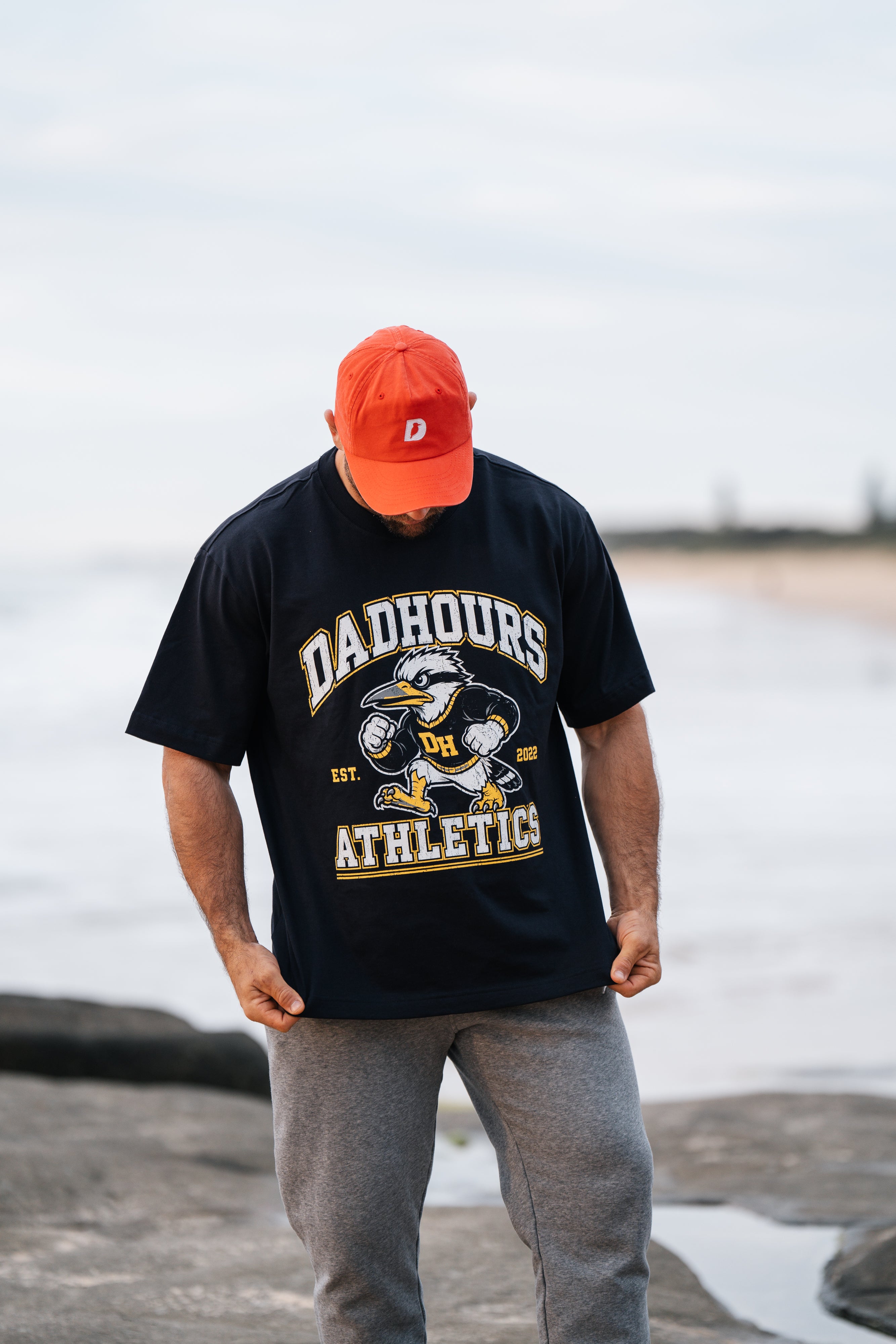 A man standing on a rocky beach wearing a black graphic t-shirt that reads 'DADHOURS ATHLETICS' with a cartoon penguin, a bright orange baseball cap, and gray sweatpants.