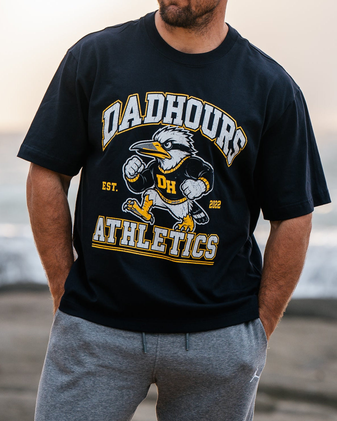 A man standing on a rocky beach wearing a black 'Dahour Athletic' t-shirt, gray sweatpants, and an orange-red baseball cap, with the ocean and bright sky in the background.