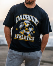 A man standing on a rocky beach wearing a black 'Dahour Athletic' t-shirt, gray sweatpants, and an orange-red baseball cap, with the ocean and bright sky in the background.