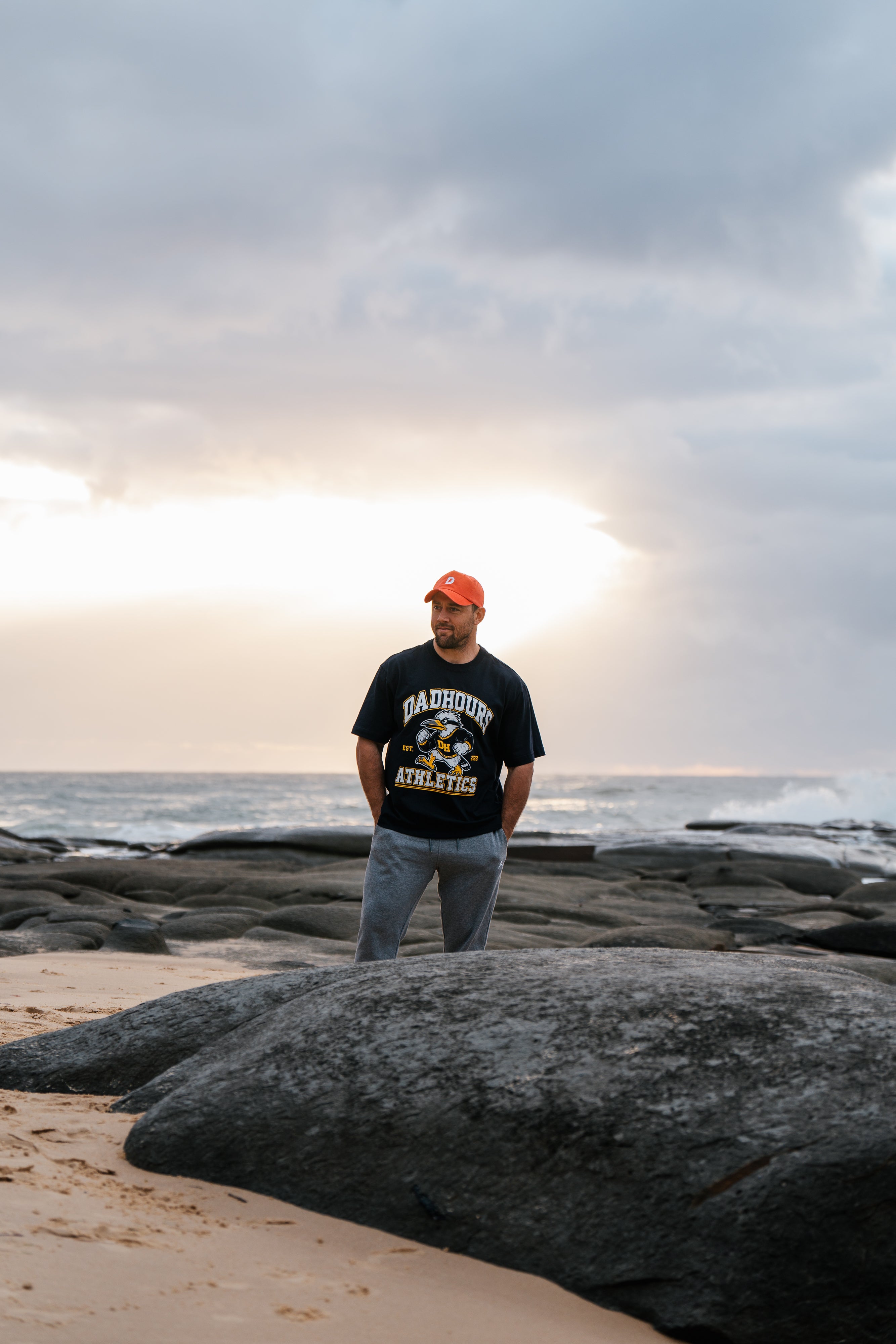 A man standing on a rocky beach wearing a black graphic t-shirt, gray pants, and an orange cap, with the ocean and a cloudy sky behind him.