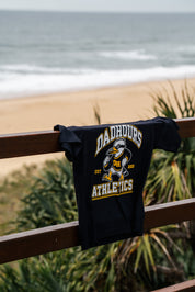 A black graphic t-shirt with 'DADROURS ATHLETICS' and a mascot design hanging from a wooden railing, with a beach and ocean in the blurred background.